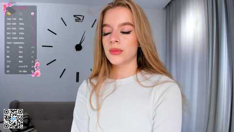 Media: A video of a young, fair-skinned woman with long blonde hair and pink lipstick, wearing a white long-sleeve top. The background features a gray wall with a large clock, a QR code, and a bed.