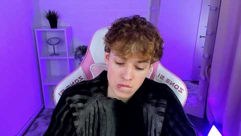 Media: Video of a young man with curly red hair, wearing a black hoodie, sitting in a white and pink gaming chair, illuminated by purple lighting in a minimalist room.