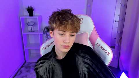 Media: Video of a young, curly-haired white male with light skin, sitting in a white gaming chair with \"Sony\" branding, wearing a black shirt. Background features a white shelf with a plant, purple lighting, and a white door.