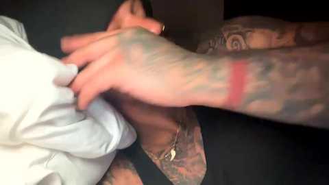 Media: Video of a close-up of a man's tattooed arm, wearing a white shirt, gripping a black shirt, in a dimly lit room.