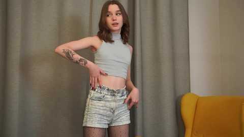 Video of a slender, young white woman with shoulder-length brown hair, wearing a light blue crop top, denim shorts, and fishnet stockings, standing confidently in front of beige curtains and a mustard-yellow chair.