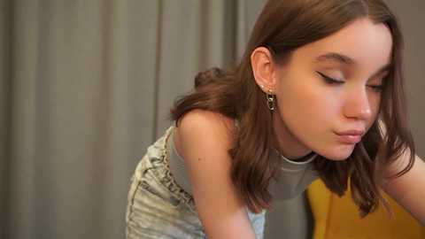 Video of a young woman with light skin, brown hair, and wearing a grey top, leaning forward with eyes closed, against a grey curtain background.