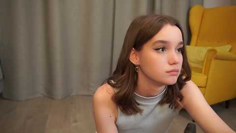 Video of a young girl with shoulder-length brown hair, wearing a gray ribbed tank top, sitting on a wooden floor. She gazes thoughtfully into the distance. Background includes a mustard-yellow armchair and gray curtains.