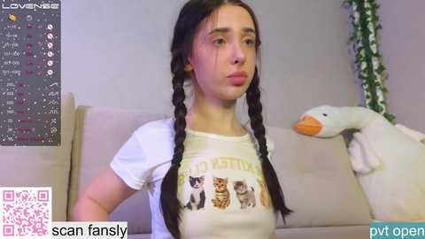 Media: A video of a young woman with long black braids, wearing a white \"LOVEME\" T-shirt, sitting on a beige sofa, with a plush duck nearby, in a casual indoor setting.