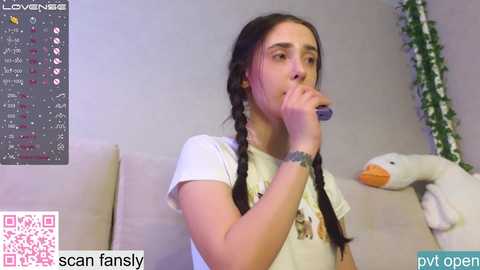 Media: Video of a young woman with long braided hair, wearing a white t-shirt, sitting on a beige couch, holding a blue toothbrush, in a cozy, indoor setting.