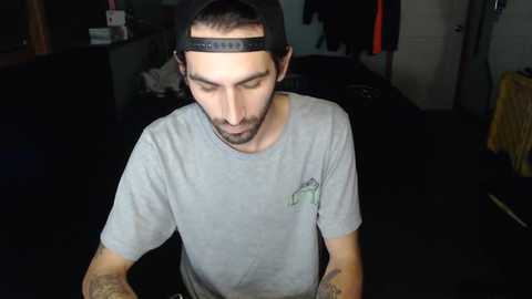 Media: Video of a man with a beard and dark hair, wearing a black cap, grey T-shirt, and tattoos, standing in a dimly lit room with dark carpet.