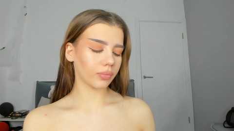 Media: Video of a young Caucasian woman with long brown hair and fair skin, wearing makeup with a bold orange eyeshadow, standing indoors in front of a white wall and door.