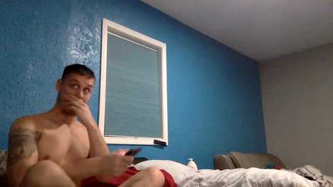 Media: Video of a shirtless, tattooed man with short dark hair, holding a phone, in a bedroom with blue walls and a window with blinds. He's sitting on a bed with white and red sheets.