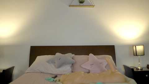 Media: Video of a minimalist bedroom with a beige bed, two white pillows, a pastel star pillow, and a plush toy, under a white wall with two hanging lamps, creating a cozy, child-friendly atmosphere.