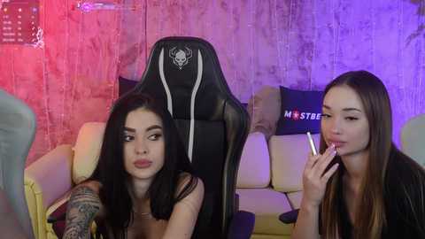 Media: Video of two young women, one with dark hair and tattoos, and one with long brown hair, sitting on gaming chairs in a dimly lit room with purple and pink lighting.