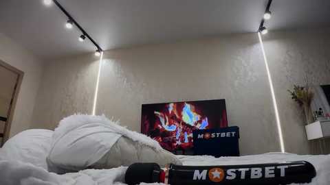 Media: Video of a minimalist bedroom with white bedding, two mounted spotlights, a flat-screen TV displaying a vibrant red and blue flame, and a black MISTBET remote control on the bed.