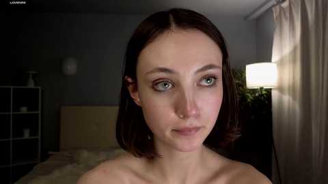 Media: Video of a young woman with fair skin, straight brown hair, and green eyes, topless, in a dimly lit bedroom with a bed, white shelves, and a lamp.