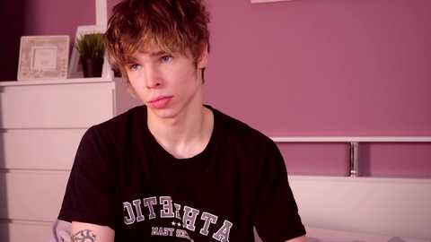 Media: Video of a young, pale-skinned, androgynous person with messy brown hair wearing a black \"JITTERY\" T-shirt, sitting on a bed in a pink room with white furniture.