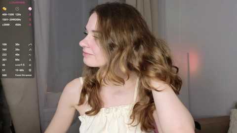 Media: Video of a young, fair-skinned woman with wavy, light brown hair, wearing a beige sleeveless top. She's in a softly lit room with beige curtains and a pink lamp.