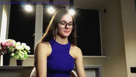 Media: Video of a young woman with long brown hair, wearing a sleeveless, form-fitting blue dress, and glasses, standing in a modern, dimly lit room with a window and a floral arrangement in the background.