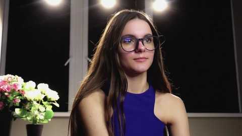 Media: Video of a young woman with long brown hair, wearing a sleeveless, dark blue dress, and black-rimmed glasses, standing indoors by a window with floral arrangement in the background.