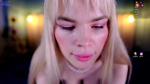 Media: Video of a fair-skinned, blonde woman with long, straight hair and pink eyeshadow, smiling softly. She wears a black top and a pearl necklace. The background is blurred with warm lighting, creating a cozy indoor ambiance.