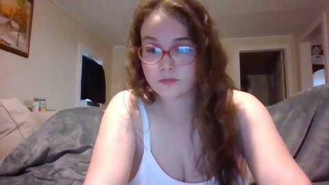 Media: Video of a young woman with long, curly brown hair and glasses, wearing a white tank top, sitting on a bed with gray bedding in a dimly lit bedroom.