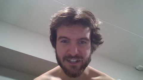 Media: Video of a smiling, bearded man with shoulder-length brown hair, standing shirtless indoors, with a plain white ceiling and wall in the background.