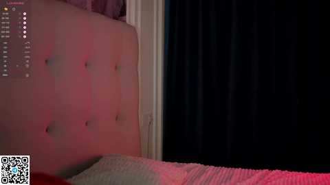 Media: A dimly lit video of a bedroom with a plush, tufted headboard, a bed with a white duvet, and dark curtains covering a window, partially illuminated by red light.