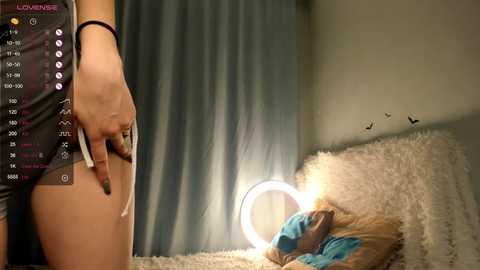 Media: A video of a woman in a black dress holding a cigarette, standing beside a bed with a blue blanket and a glowing ring light. The room features a soft, beige wall and a window with blue curtains.