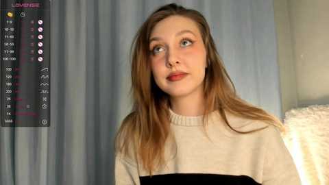 Media: Video of a young woman with light skin, long brown hair, wearing a cream-colored sweater, looking contemplative. Background includes a gray curtain and a white textured pillow.