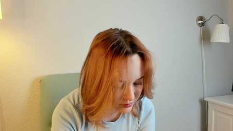 Media: Video of a Caucasian woman with shoulder-length, light brown hair, wearing a light blue sweater, sitting on a light green chair against a plain light gray wall, with a white lamp on the right.