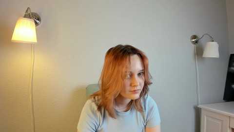 Media: Video of a young woman with shoulder-length, reddish-brown hair, wearing a light blue T-shirt, sitting in a room with two wall-mounted lamps, one lit, on light gray walls.