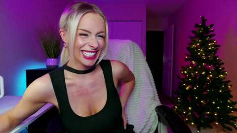 Media: Video of a smiling, fair-skinned blonde woman in a black sleeveless top, sitting in a chair, with a lit Christmas tree in the background under purple lighting.