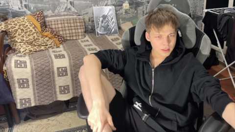 Media: Video of a young Caucasian man with light skin, short brown hair, and a lean physique, seated in a gaming chair, wearing black hoodie and shorts, surrounded by gaming accessories and a leopard-print bedspread.