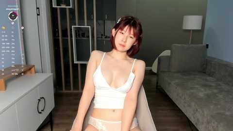 Media: A young Asian woman with red hair sits in a modern living room, wearing a white lace bra and panties, looking downcast.