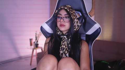 Media: Video of a woman with long black hair, wearing glasses, leopard print scarf, and black top, sitting in a gaming chair in a dimly lit room with a wooden wall and a lamp.