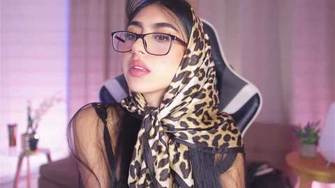 Media: Video of a young woman with medium skin tone, black hair, wearing leopard-print scarf, glasses, and a sheer black top, seated in a gaming chair in a dimly lit room.