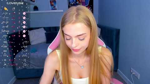 Media: A video of a young woman with long blonde hair, wearing a white tank top, sitting on a pink gaming chair in a blue-themed bedroom.