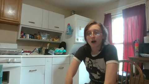 Media: A video of a young woman with glasses, short brown hair, and a black t-shirt, leaning over a kitchen sink. The kitchen is cluttered with white cabinets, a stove, and a sink, with red curtains in the background.