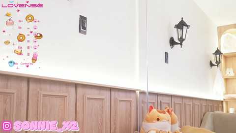 Media: Video of a modern, minimalist kitchen with white walls, wooden wainscoting, and a glass cabinet. A \"LOVENSE\" logo and a cat-themed sticker are on the wall, along with a black lantern.