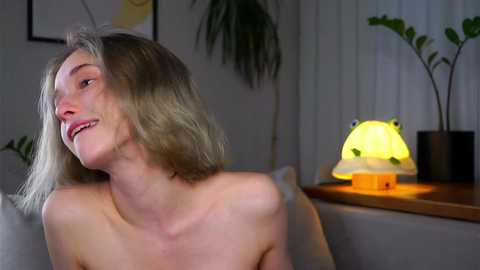 Video of a smiling, topless Caucasian woman with shoulder-length blonde hair, seated in a dimly lit room with a green plant and a lamp with a mushroom-shaped shade.