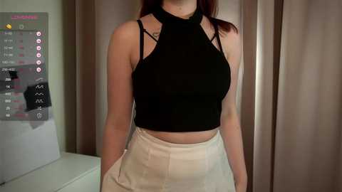 Media: Video of a fair-skinned woman with red hair wearing a black halter top and high-waisted beige skirt, standing indoors with beige curtains in the background.