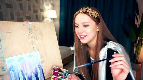 Media: Video of a young, fair-skinned woman with long, straight brown hair adorned with a gold headband, painting in a studio with blue curtains, a beige couch, and abstract artwork.