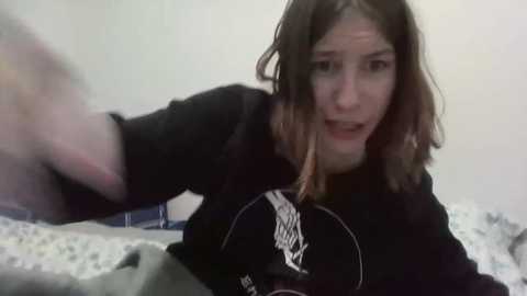 Media: A blurry video of a young woman with shoulder-length, wavy brown hair, wearing a black hoodie, lying on a bed with a white and blue patterned blanket. She appears to be startled, her mouth open.