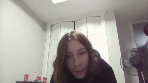 Media: Video of a young woman with long brown hair, wearing a black jacket, in a bathroom with white walls, a cabinet, and a standing fan. Red bottles on a countertop in the foreground.