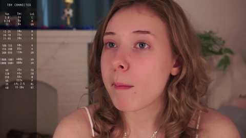 Media: Video of a young Caucasian woman with wavy blonde hair, light skin, and blue eyes, wearing a white tank top, looking introspective. Background includes a TV with a sports score, a fireplace, and a potted plant.