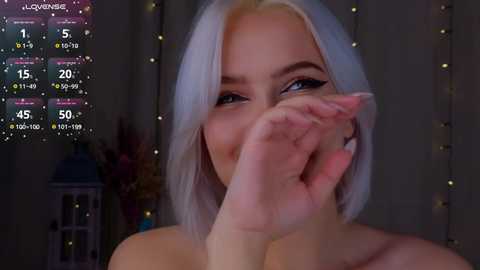 Media: Video of a young woman with platinum blonde hair, wearing black eyeliner, covering her mouth with her hand, in a dimly lit room with a digital clock and string lights in the background.