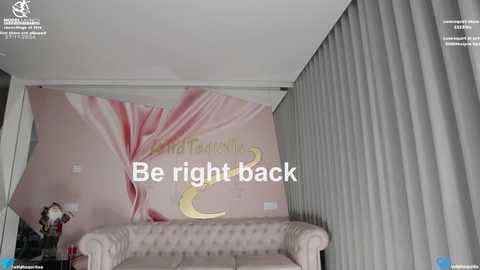 Media: Video of a modern living room with a pink and white floral mural, a beige tufted sofa, and grey vertical blinds. Text overlay reads \"Be right back.\