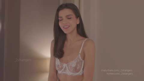 Media: Video of a slender, fair-skinned woman with long, straight dark hair, wearing a sheer, white lace bra, smiling softly, standing against a neutral-toned background.