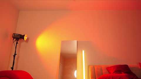 Media: Video of a modern bedroom bathed in warm, red-orange light. A tall, cylindrical lamp casts a soft glow on a white wall and a large, vertical mirror. A bed with red bedding is partially visible in the background.