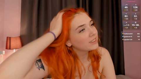 Media: Video of a fair-skinned woman with long, vibrant red hair, smiling while a hand massages her head. She has a tattoo on her upper arm. Background includes a dark curtain and a bedside lamp.