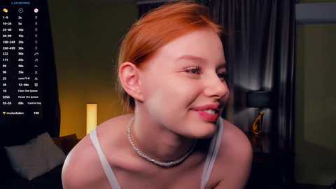 Media: A video of a young, smiling, red-haired woman with fair skin, wearing a white tank top and pearl necklace, in a dimly-lit room with a lit lamp and curtains.