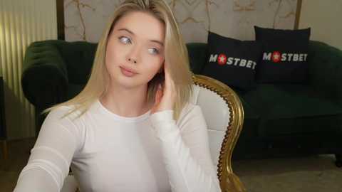 Media: Video of a young Caucasian woman with blonde hair, wearing a white long-sleeve top, sitting indoors, with a plush green couch and decorative pillows in the background.