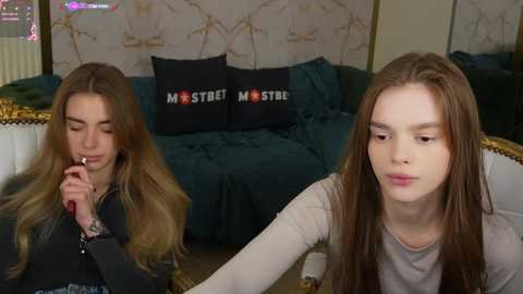 Media: Video of two young women with long brown hair, one smoking, sitting on a plush green sofa with gold accents. Background shows a wall with a pattern and a window with curtains.